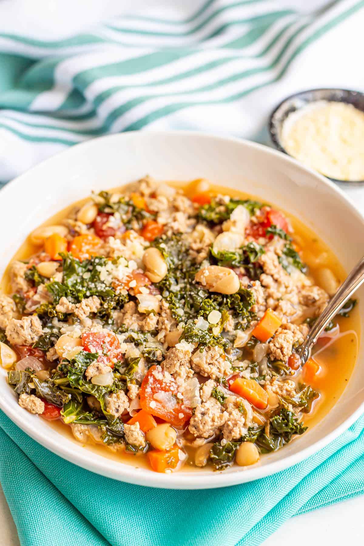 A spoon resting in a white bowl of soup with ground turkey, kale, carrots, tomatoes and white beans.