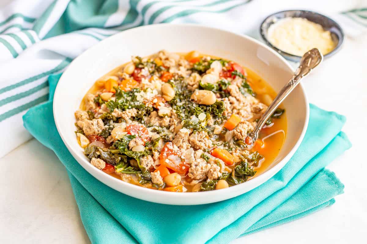 A low white bowl filled with a ground turkey soup with kale and white beans with a spoon resting in it.