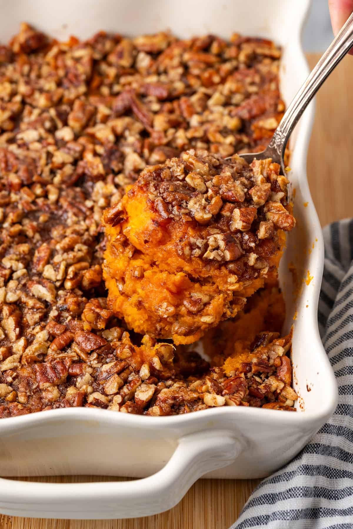 A spoon holding up a scoop of bourbon sweet potato casserole with a nutty streusel topping from a white casserole dish.