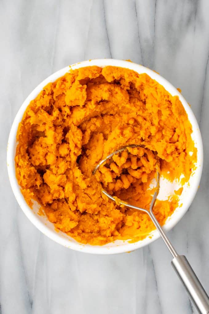 A potato masher resting in a white bowl of mashed sweet potatoes with bourbon and seasonings.