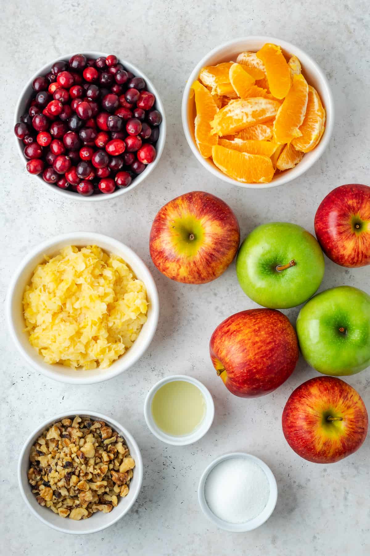 Apples, cranberries, oranges and other ingredients for a winter fruit salad laid out in separate bowls on a counter.