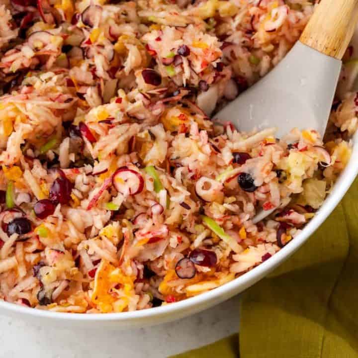 A spatula resting in a large white bowl of a cranberry apple salad with oranges and walnuts.
