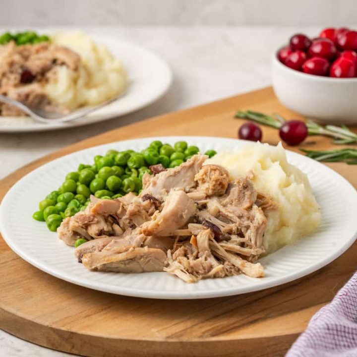 Cranberry Dijon chicken served alongside mashed potatoes and peas on a white dinner plate with a bowl of cranberries in the background.