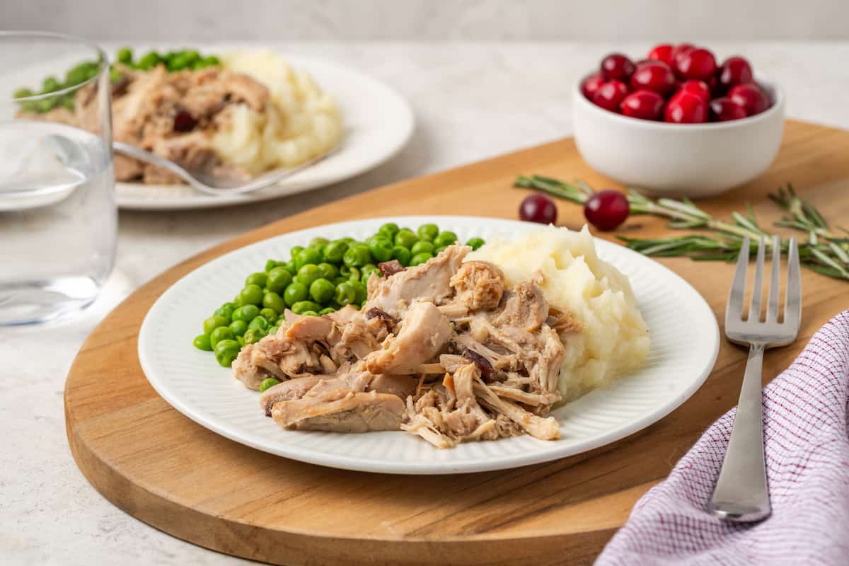 Cranberry Dijon chicken served alongside mashed potatoes and peas on a white dinner plate with a bowl of cranberries in the background.
