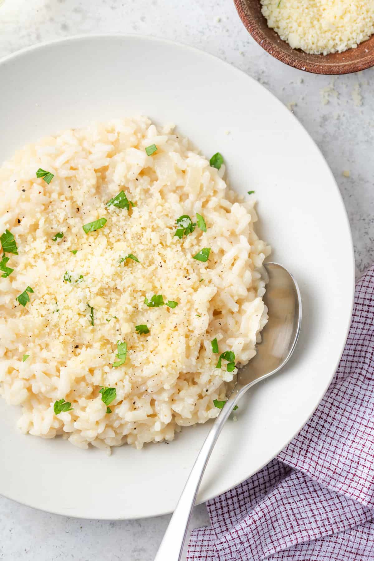 A spoon resting in a white bowl of risotto topped with Parmesan cheese, black pepper and chopped fresh parsley.