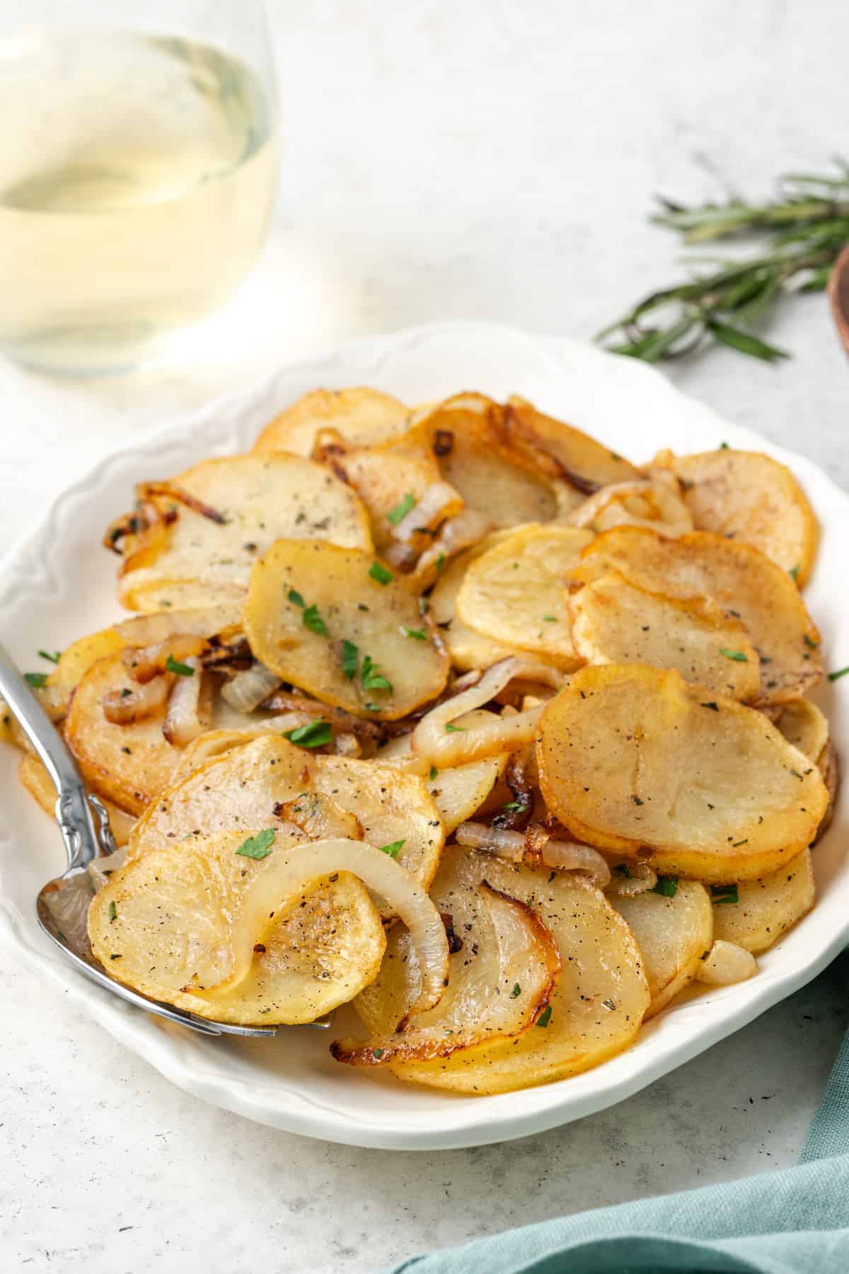 A white serving dish of Potatoes Lyonnaise with a glass of wine and some fresh herbs in the background.