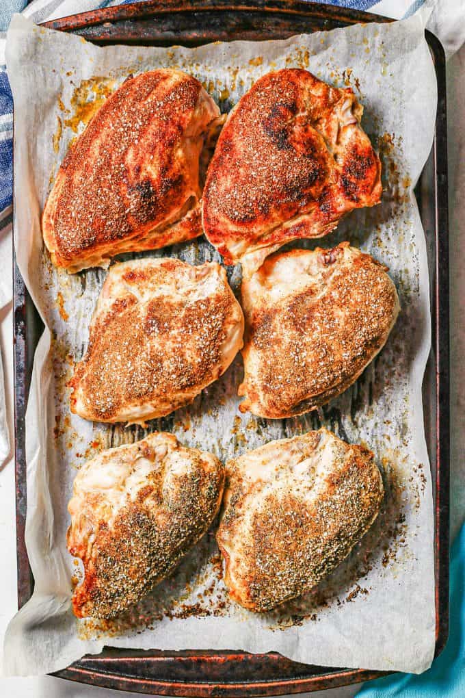 Six split chicken breasts on a parchment paper lined baking sheet with three different seasoning mixes after roasting in the oven.