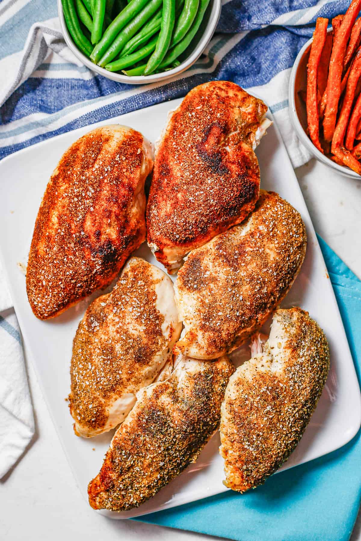 A platter of six roasted chicken breasts with different seasonings alongside bowls of green beans and sweet potato fries.