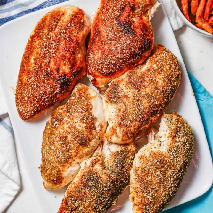 Close up of six roasted chicken breasts with different seasonings on a white platter.