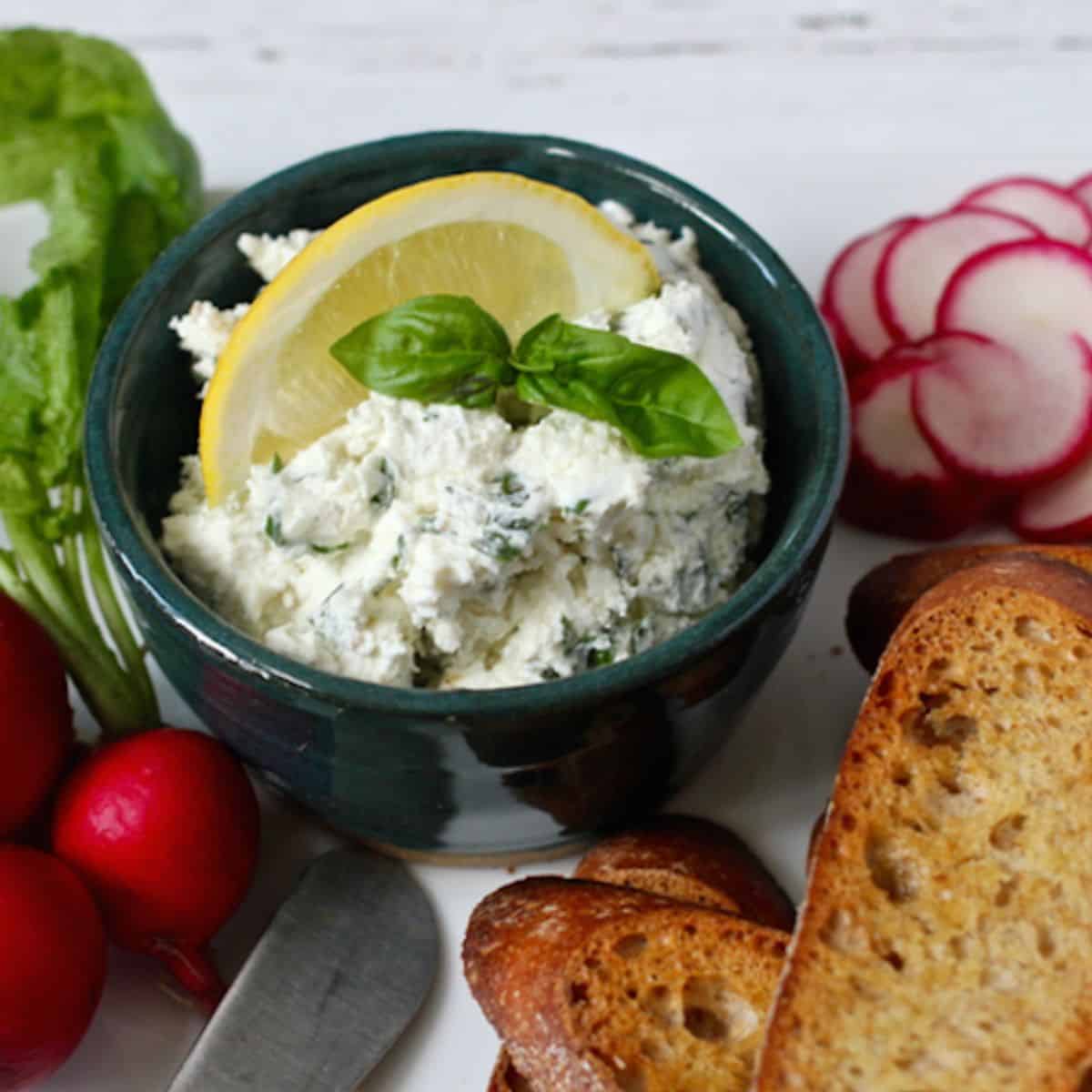 A cheese herb spread in a small green bowl with a mint sprig and lemon slice on top and sliced radishes and baguette slices nearby.