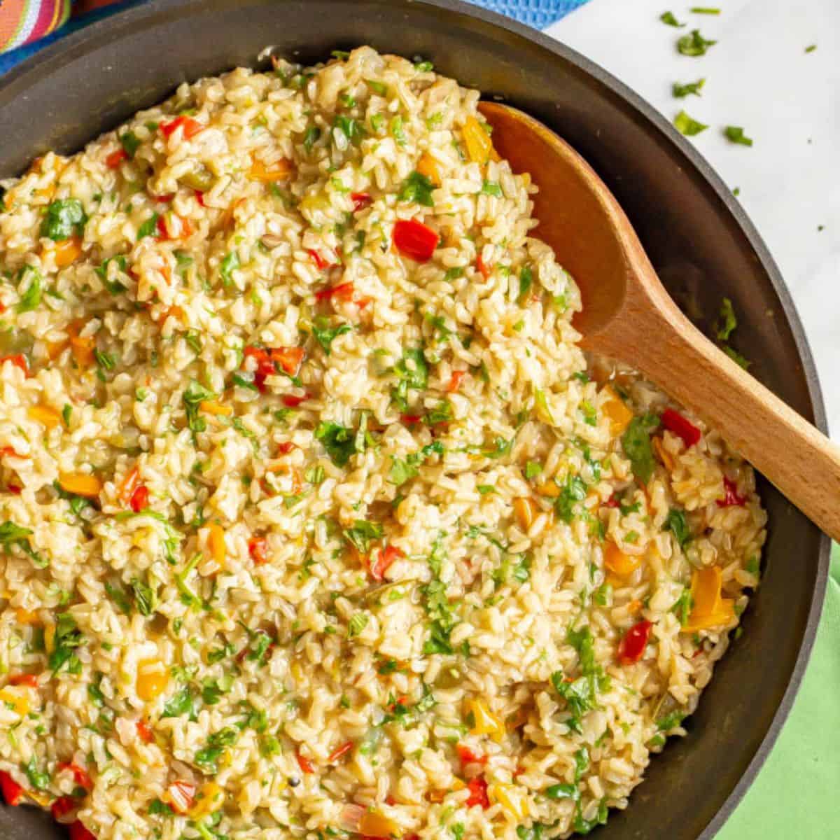 A skillet of a colorful brown rice with peppers and cilantro and a wooden spoon resting in the pan.
