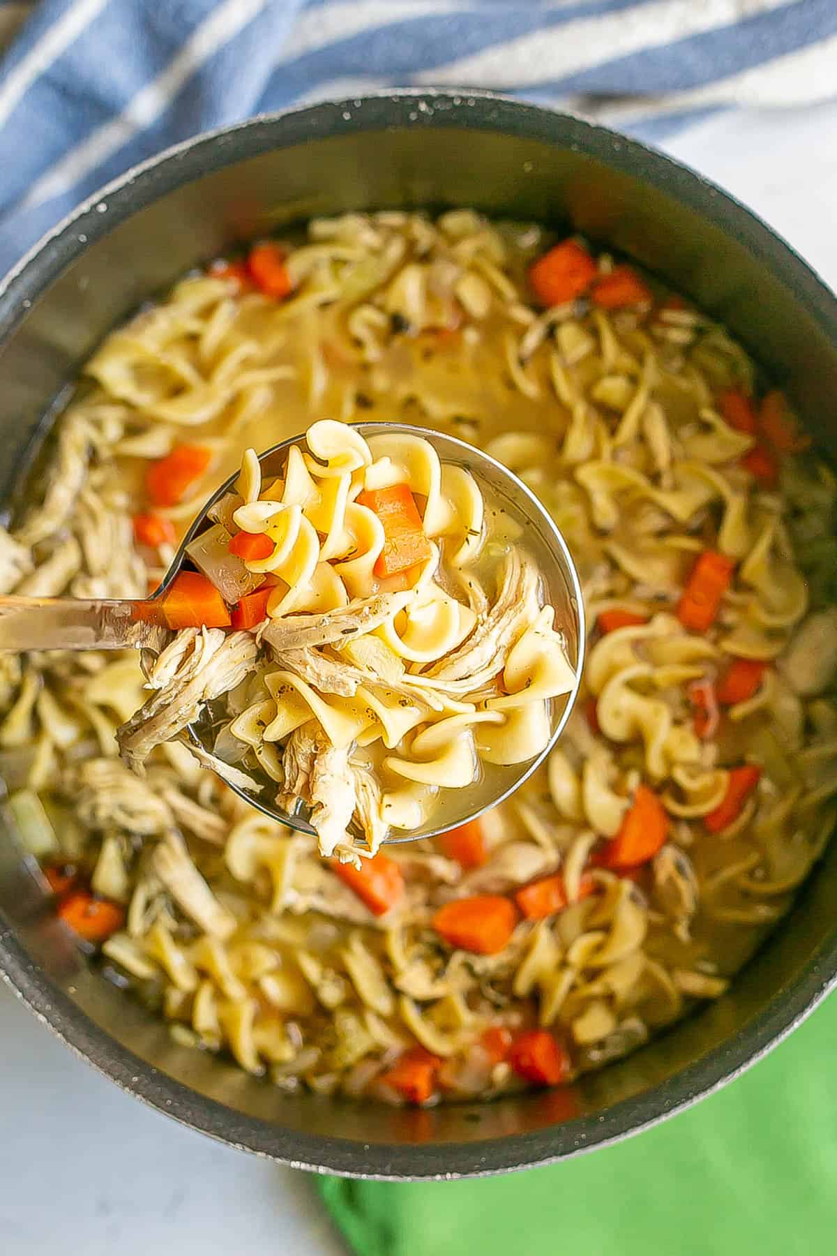 A ladle of chicken noodle soup scooped out from a large dark pot.