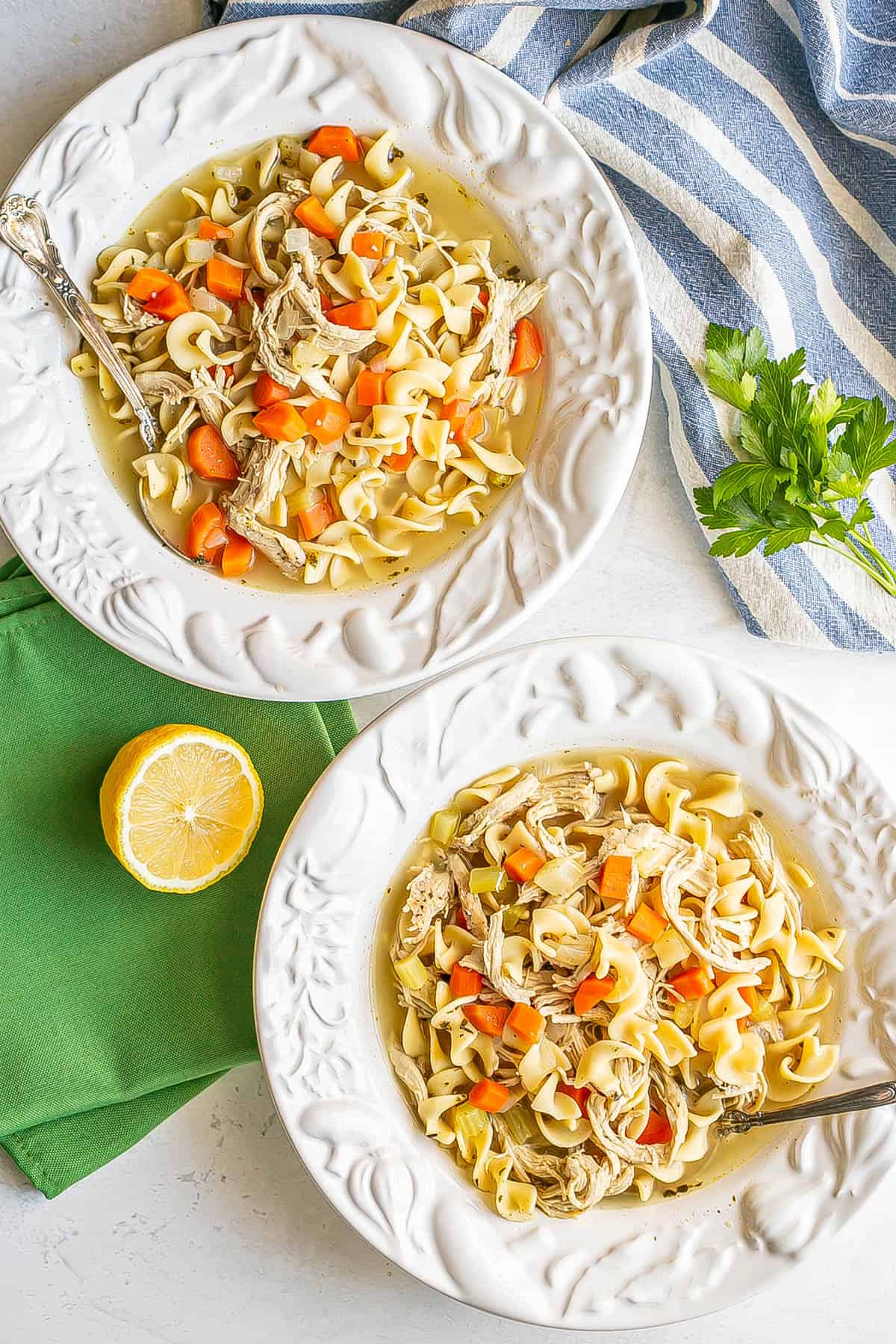 Two bowls of chicken noodle soup with spoons resting in it and parsley and lemon to the side.