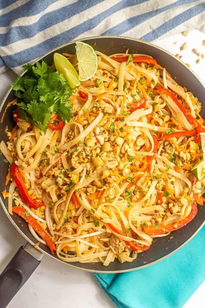 Homemade easy chicken pad Thai in a large dark skillet with chopped peanuts on top and lime wedges and cilantro to the side.