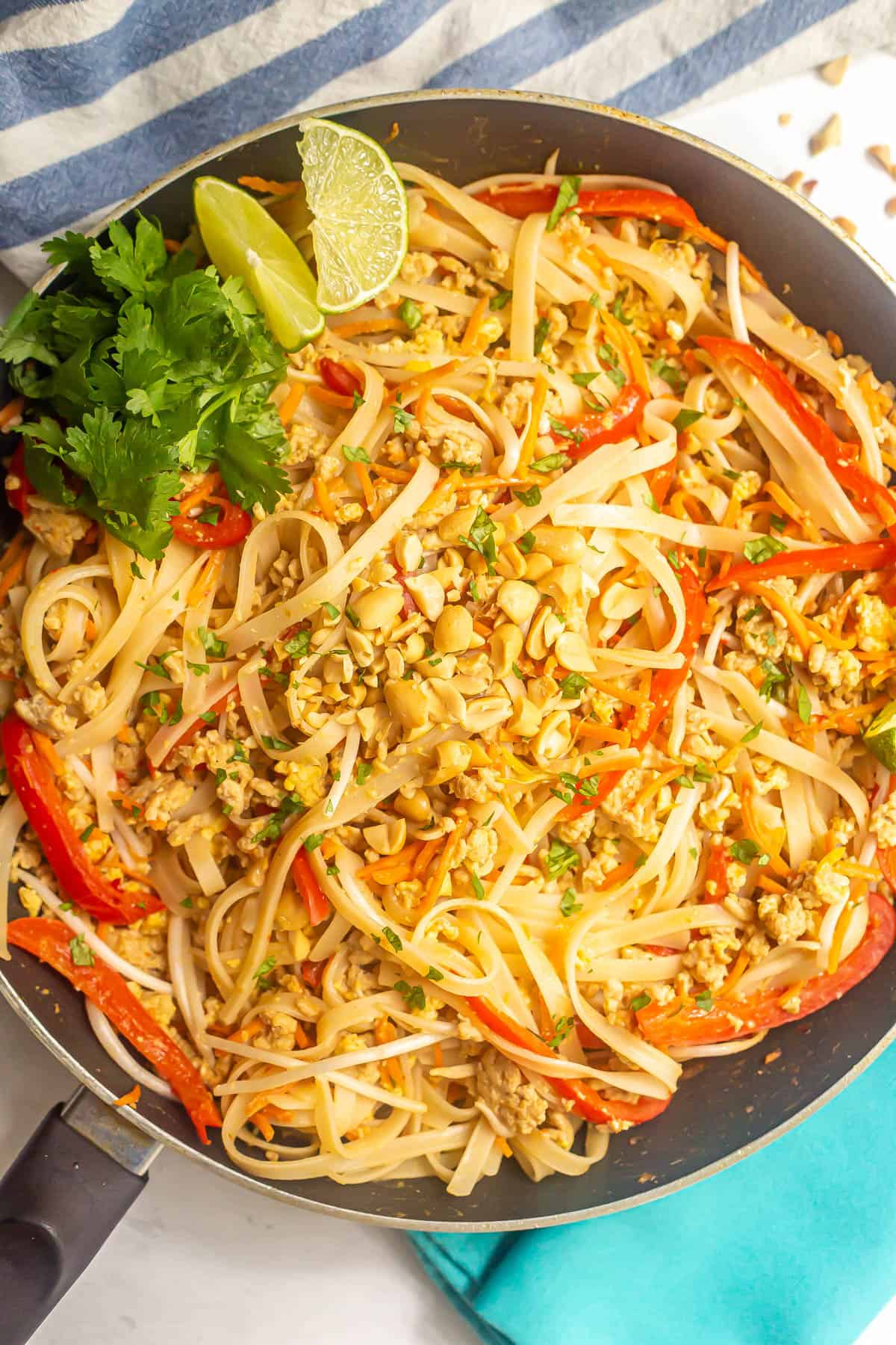 Close up of homemade easy chicken pad Thai in a large dark skillet with chopped peanuts on top and lime wedges and cilantro to the side.