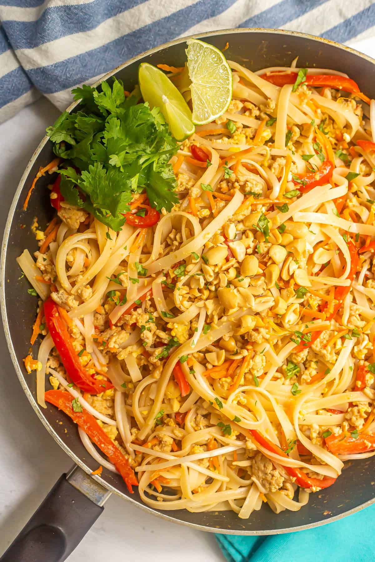 Chicken pad Thai in a large dark skillet with lime wedges and cilantro.