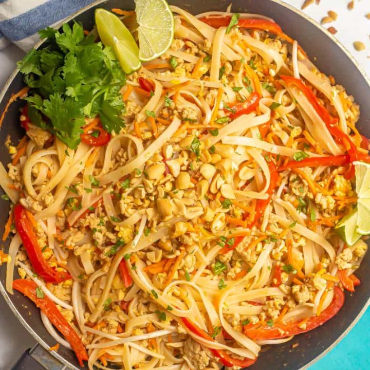 Easy pad Thai with ground chicken, noodles and red peppers in a large dark skillet with peanuts on top and lime wedges and cilantro to the side.