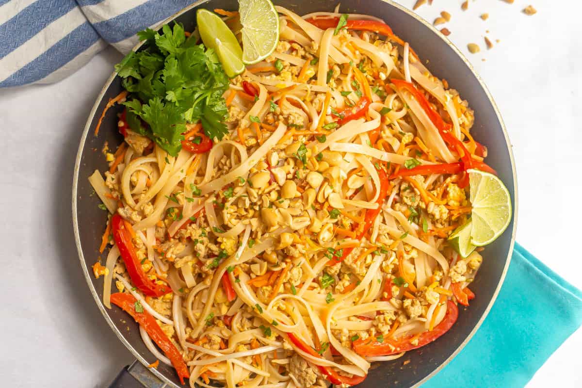 Easy pad Thai with ground chicken, noodles and red peppers in a large dark skillet with peanuts on top and lime wedges and cilantro to the side.