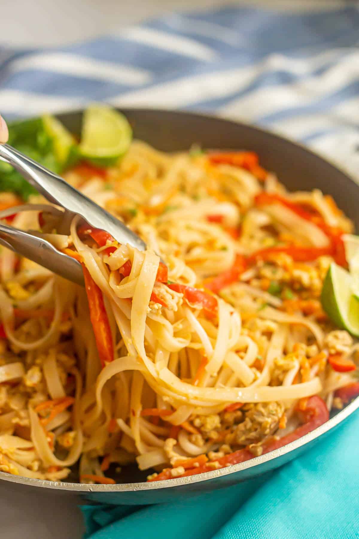 Tongs picking up a scoop of easy chicken pad Thai noodles and veggies from a large dark skillet.