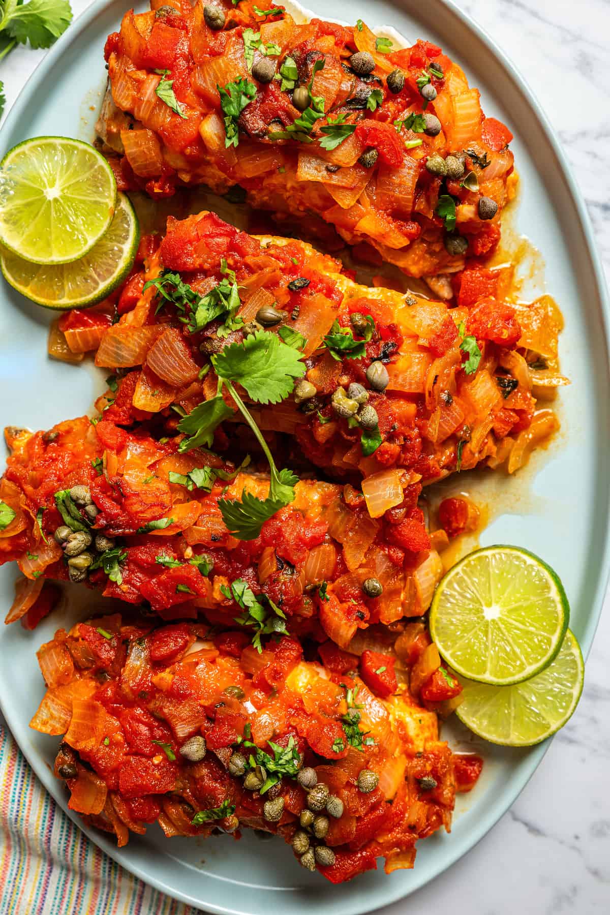 Baked grouper veracruz served on an oval serving platter with fresh herbs and lime slices.