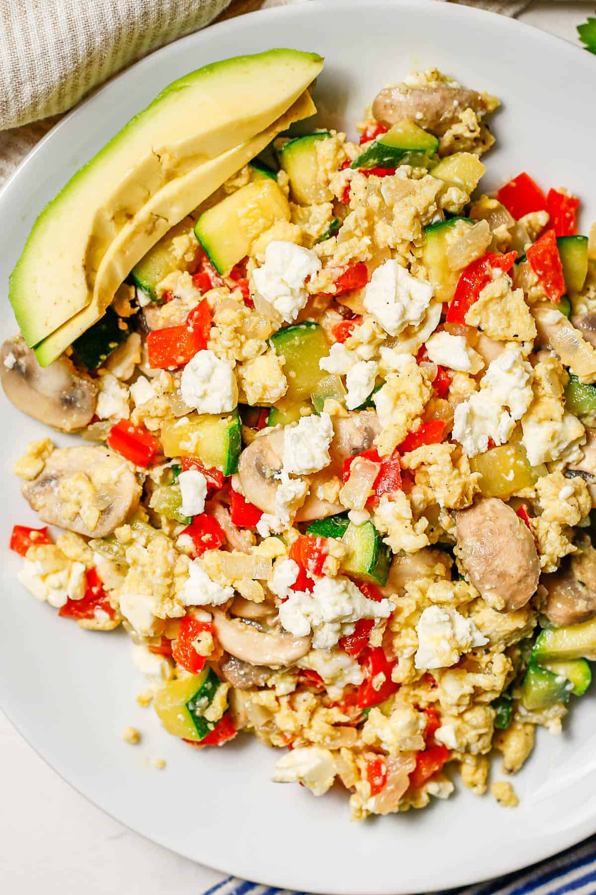 Close up of a white plate with a scrambled egg vegetable mixture with feta cheese on top and avocado slices to the side.