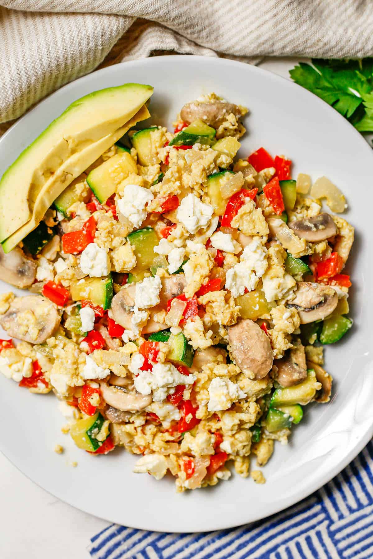 A white plate with a scrambled egg vegetable mixture with feta cheese on top and avocado slices to the side.