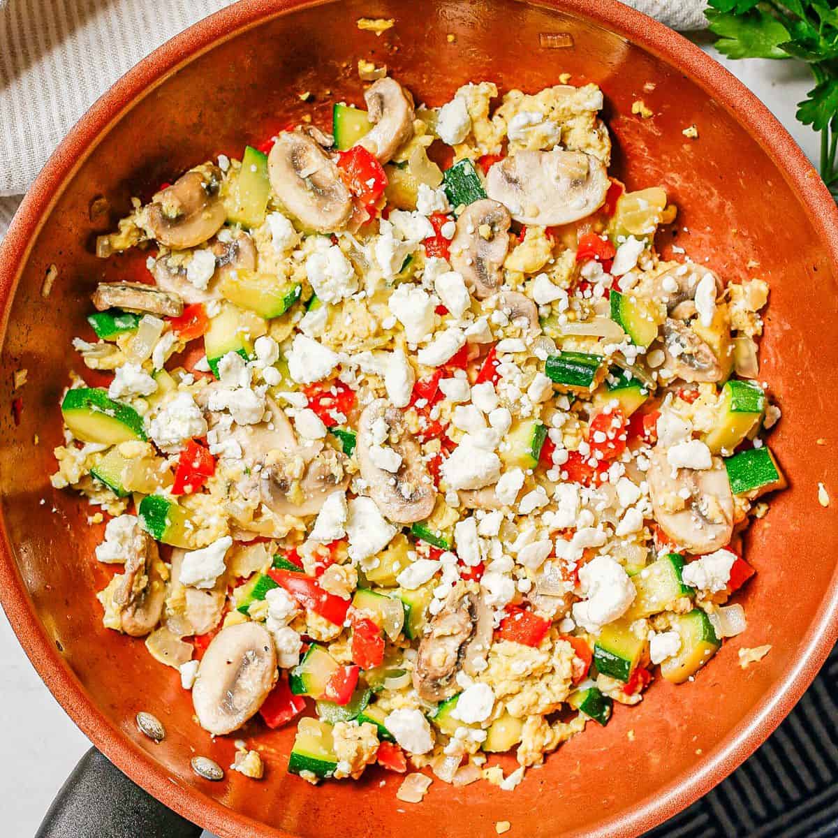 Close up of a copper skillet with an egg scramble with vegetables topped with feta cheese.