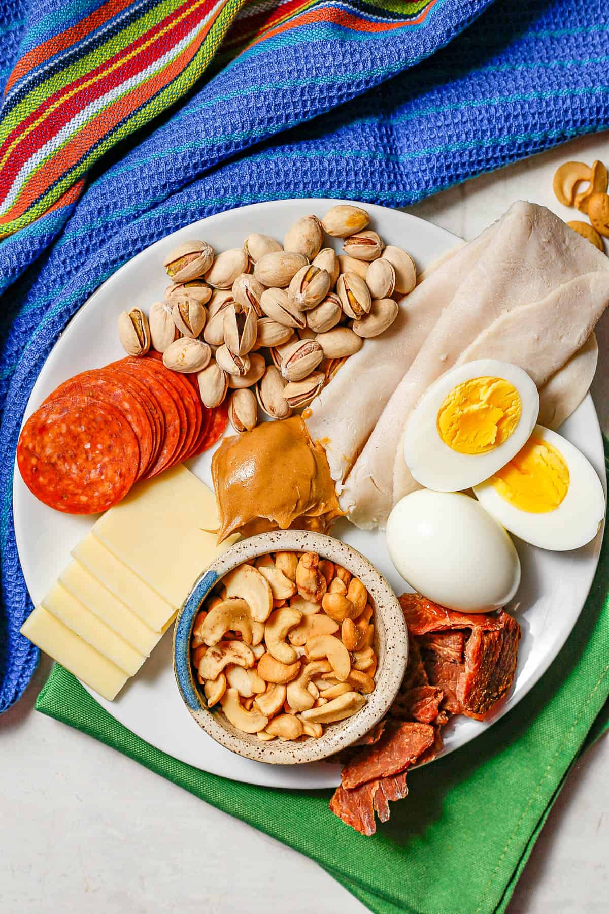 A white plate with a variety of healthy high protein snacks, including cheese, pepperoni, nuts, turkey, hard boiled eggs and peanut butter.