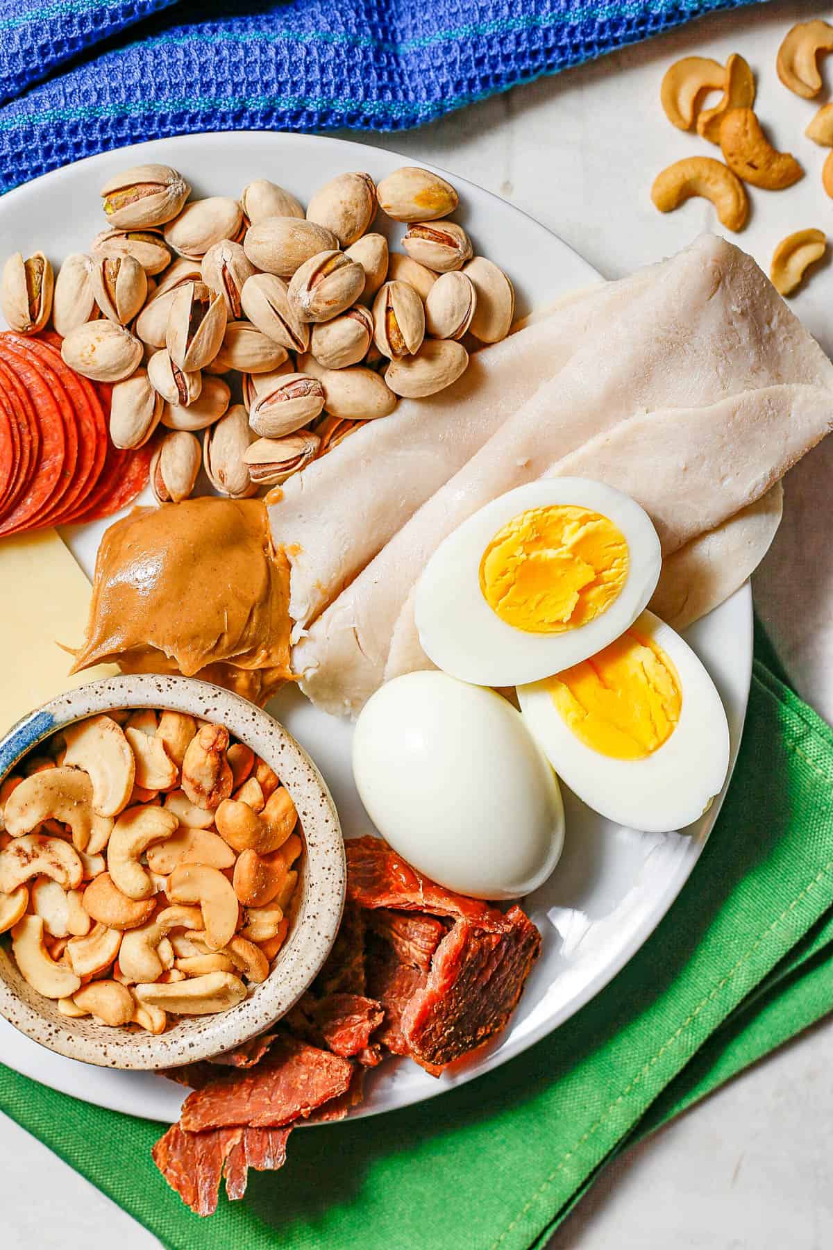 Close up of a white plate with a variety of healthy high protein snacks, including cheese, pepperoni, nuts, turkey, hard boiled eggs and peanut butter.