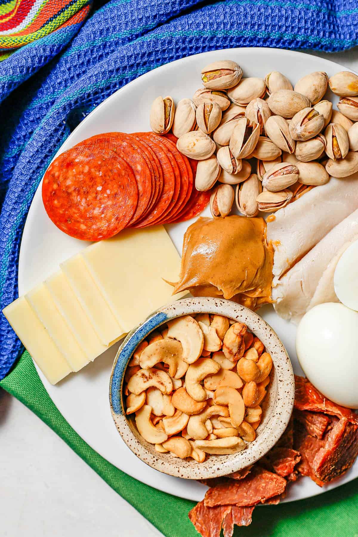 Nuts, peanut butter, pepperoni and cheese slices on a white plate set on green napkins.