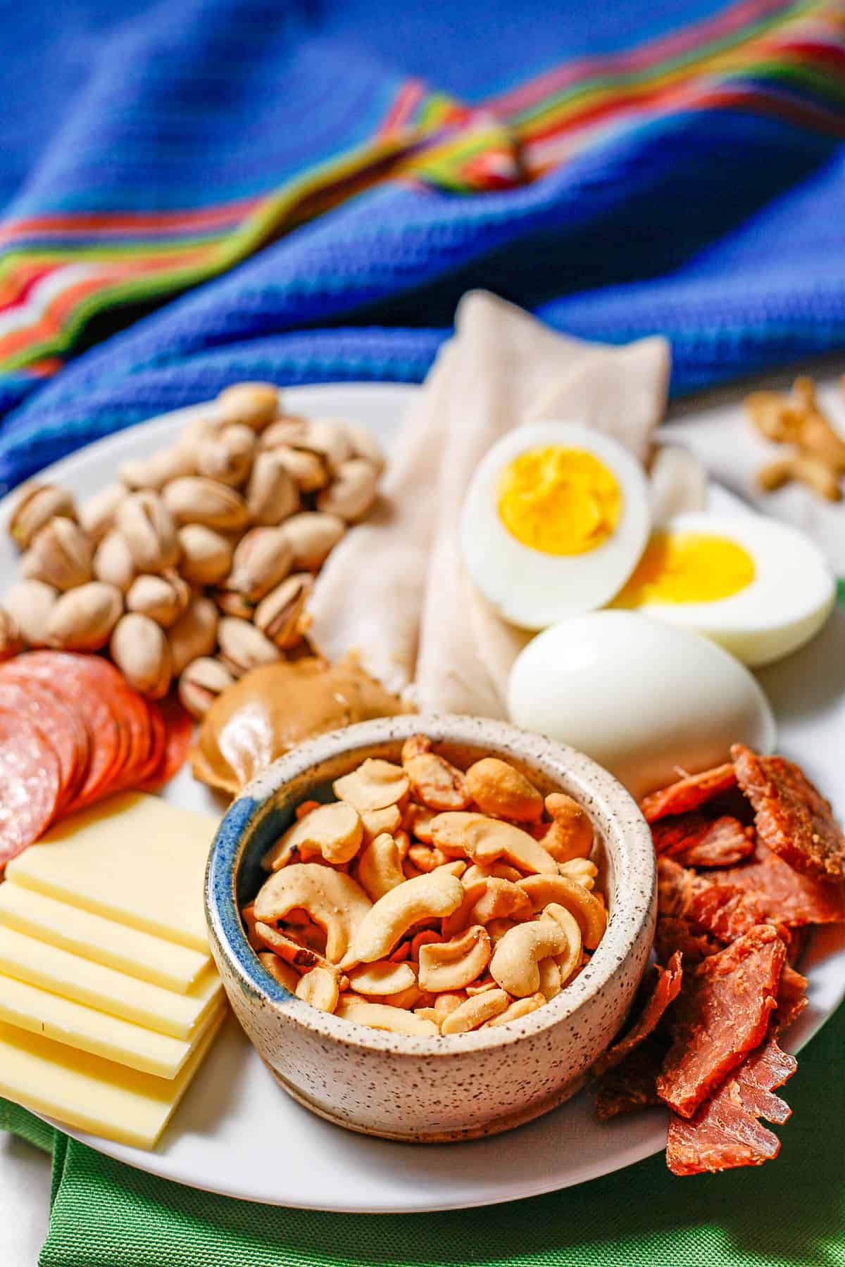 A bowl of cashews on a white plate with other high protein healthy snacks.