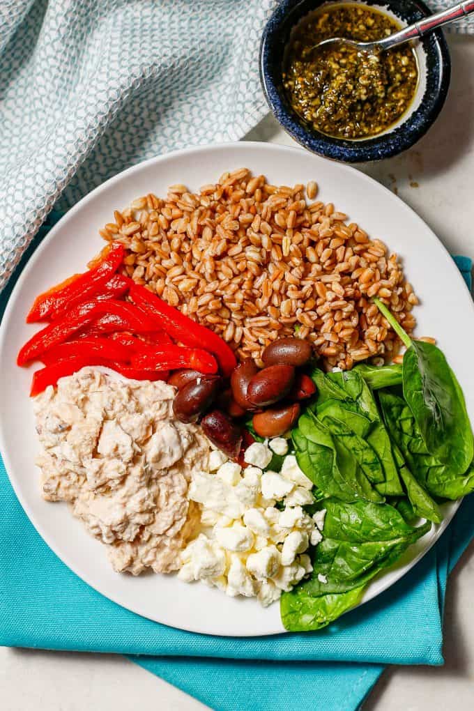A white dinner plate arranged with a creamy canned salmon mixture, roasted red peppers, farro, spinach, feta and olives with a bowl of pesto to the side.