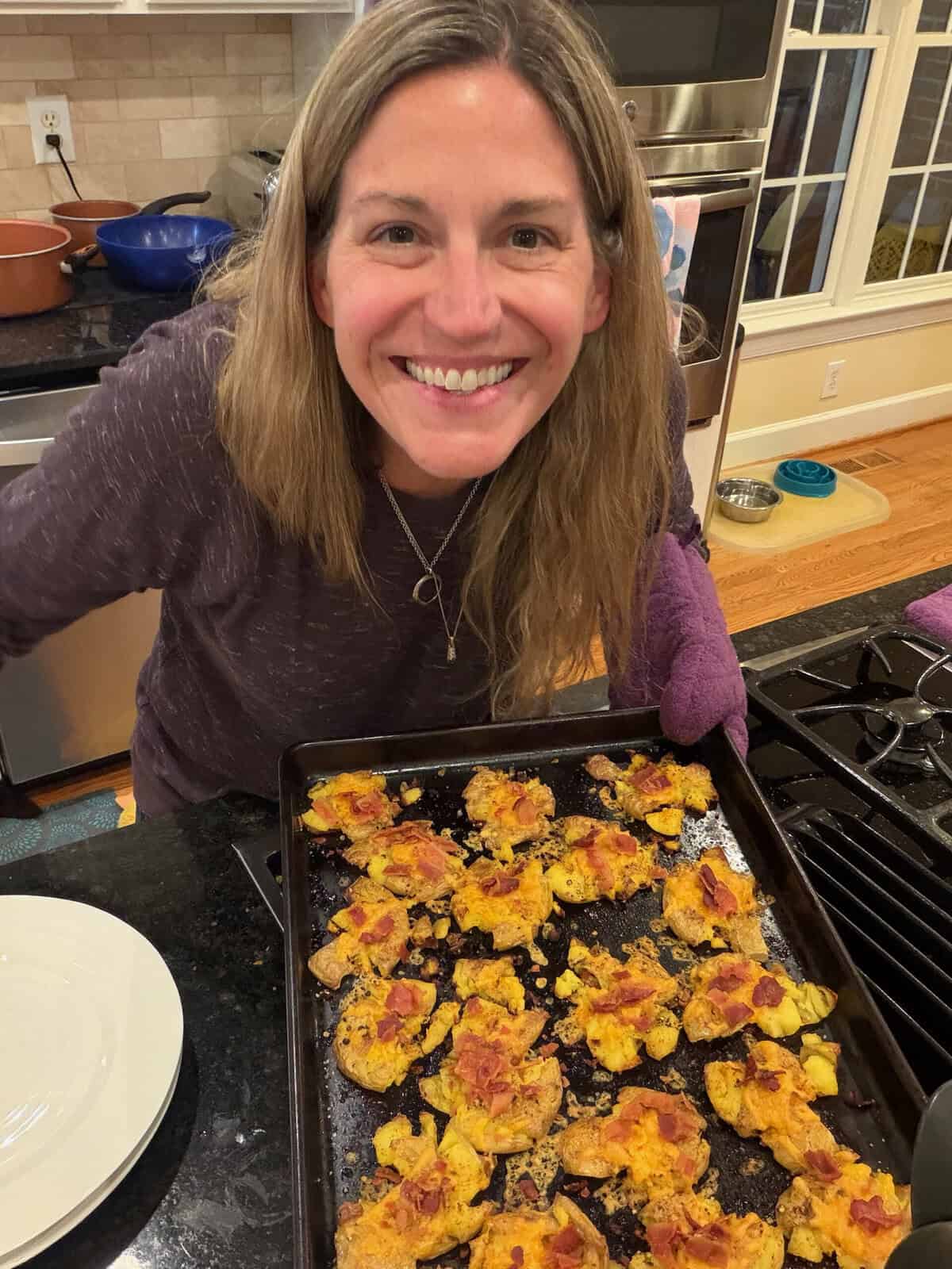 Kathryn in her kitchen holding a baking sheet filled with smashed potatoes with cheese and bacon on top.
