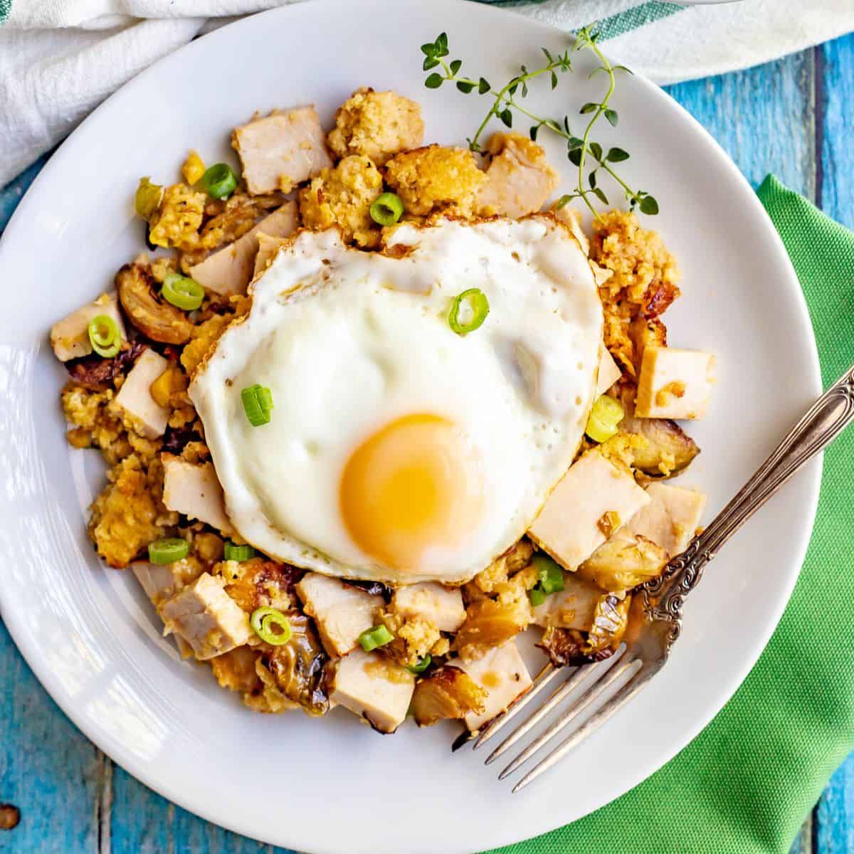 Close up of a fried egg on top of a breakfast hash made with Thanksgiving leftovers on a white plate with a fork resting on the side.