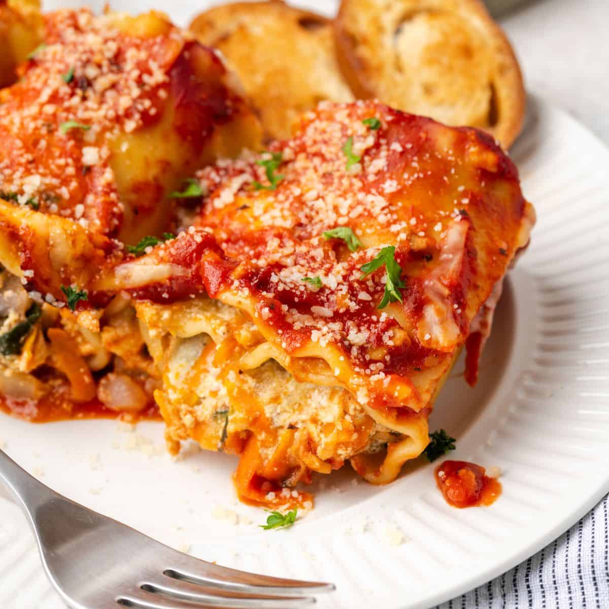 Close up of baked vegetable lasagna rolls on a white plate with cheese and chopped parsley sprinkled on top.