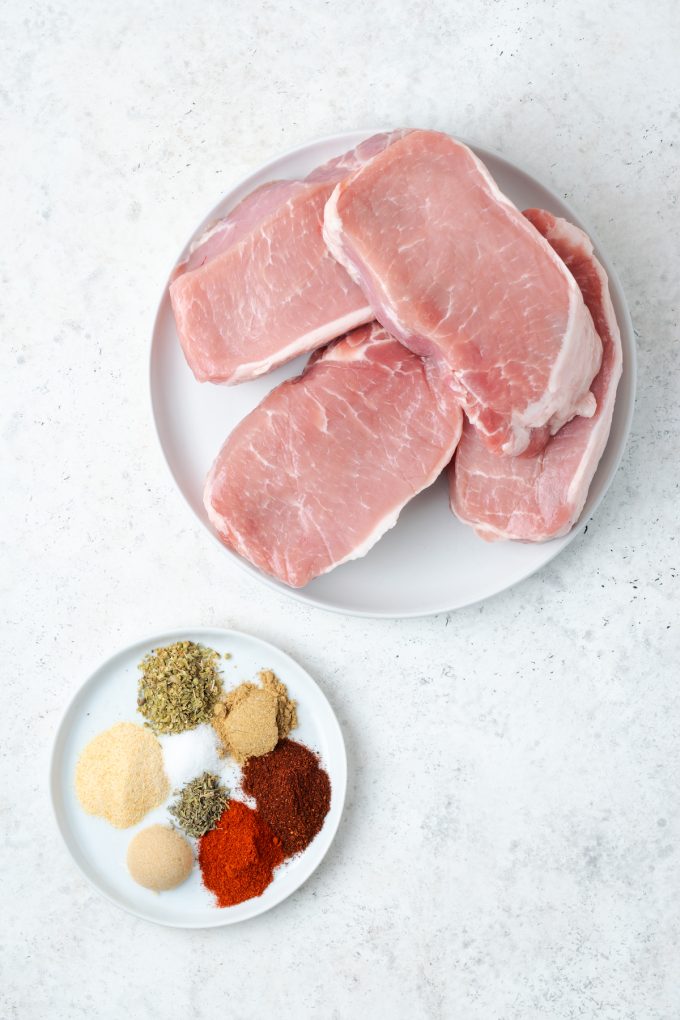 Four pork chops on a plate with a separate small plate of a seasoning mix in separate piles.