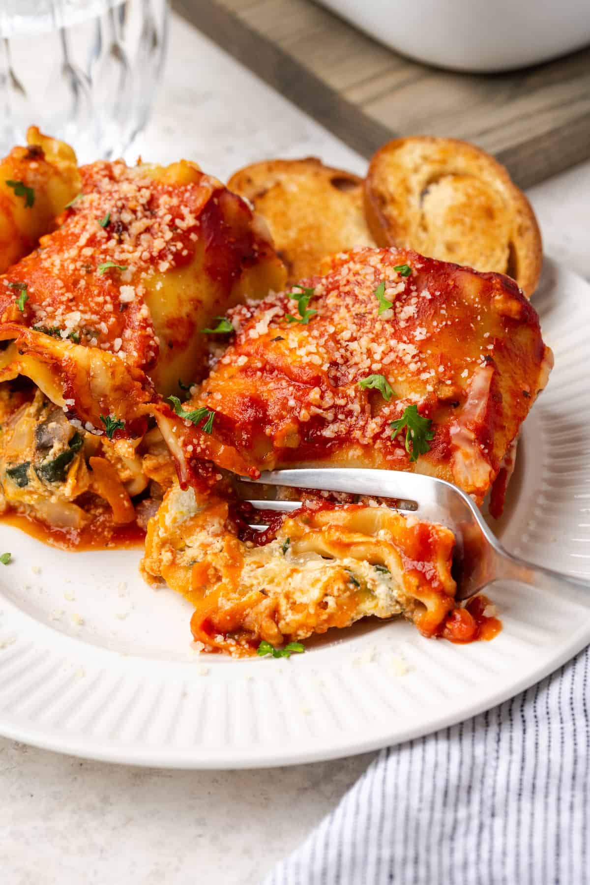 A fork cutting into a baked cheesy vegetable lasagna roll up on a white plate.