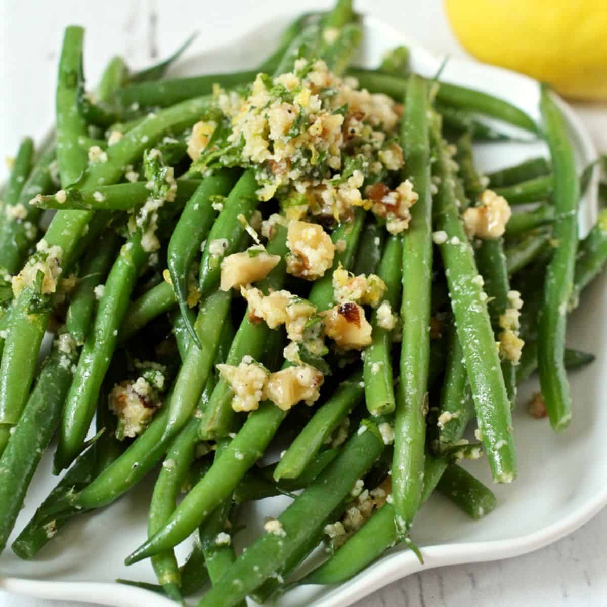 Close up of a white plate with a pile of green beans topped with a gremolata mixture of walnuts and herbs and lemon.