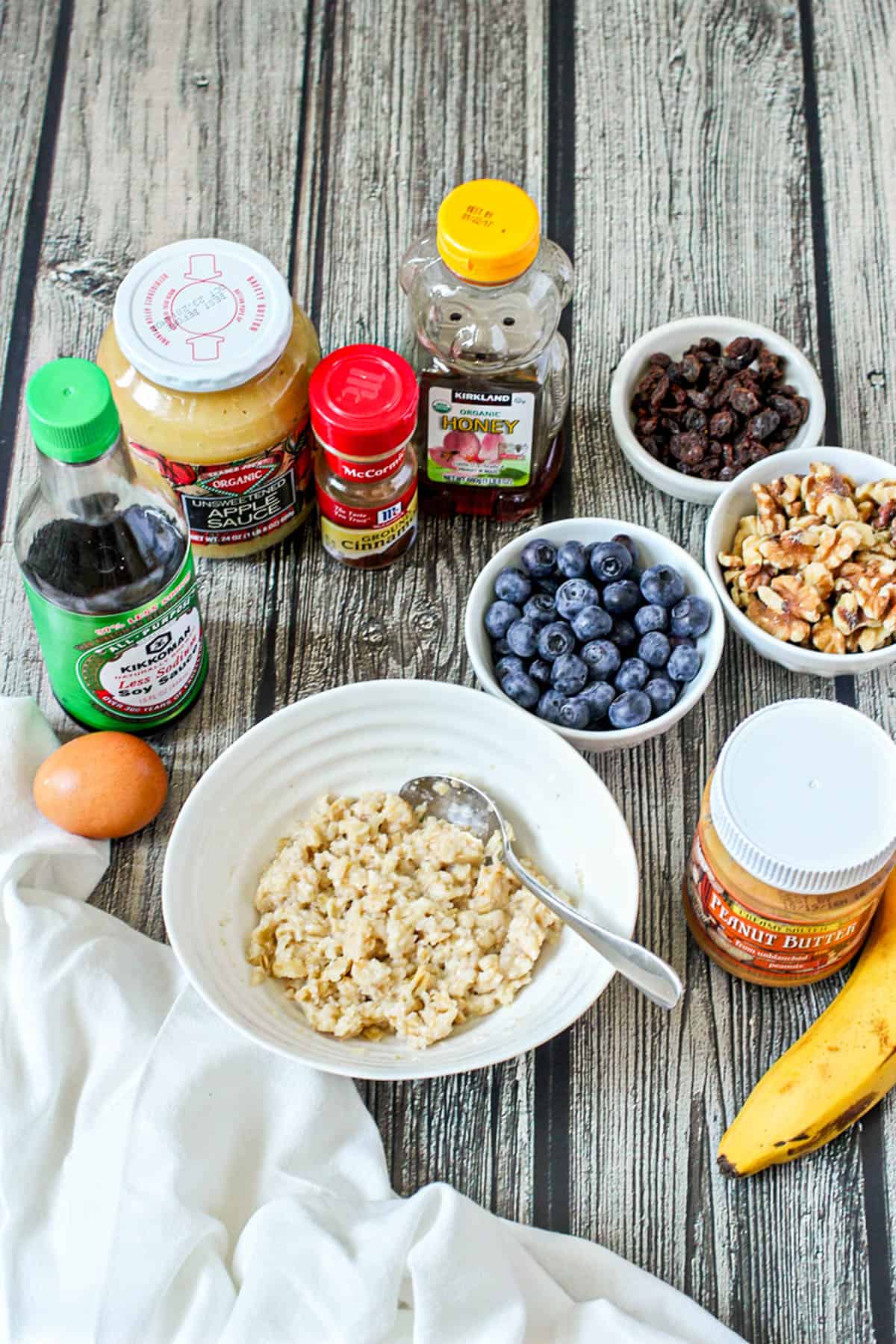 A bowl of oatmeal surrounded by toppings including blueberries, applesauce, peanut butter and banana.