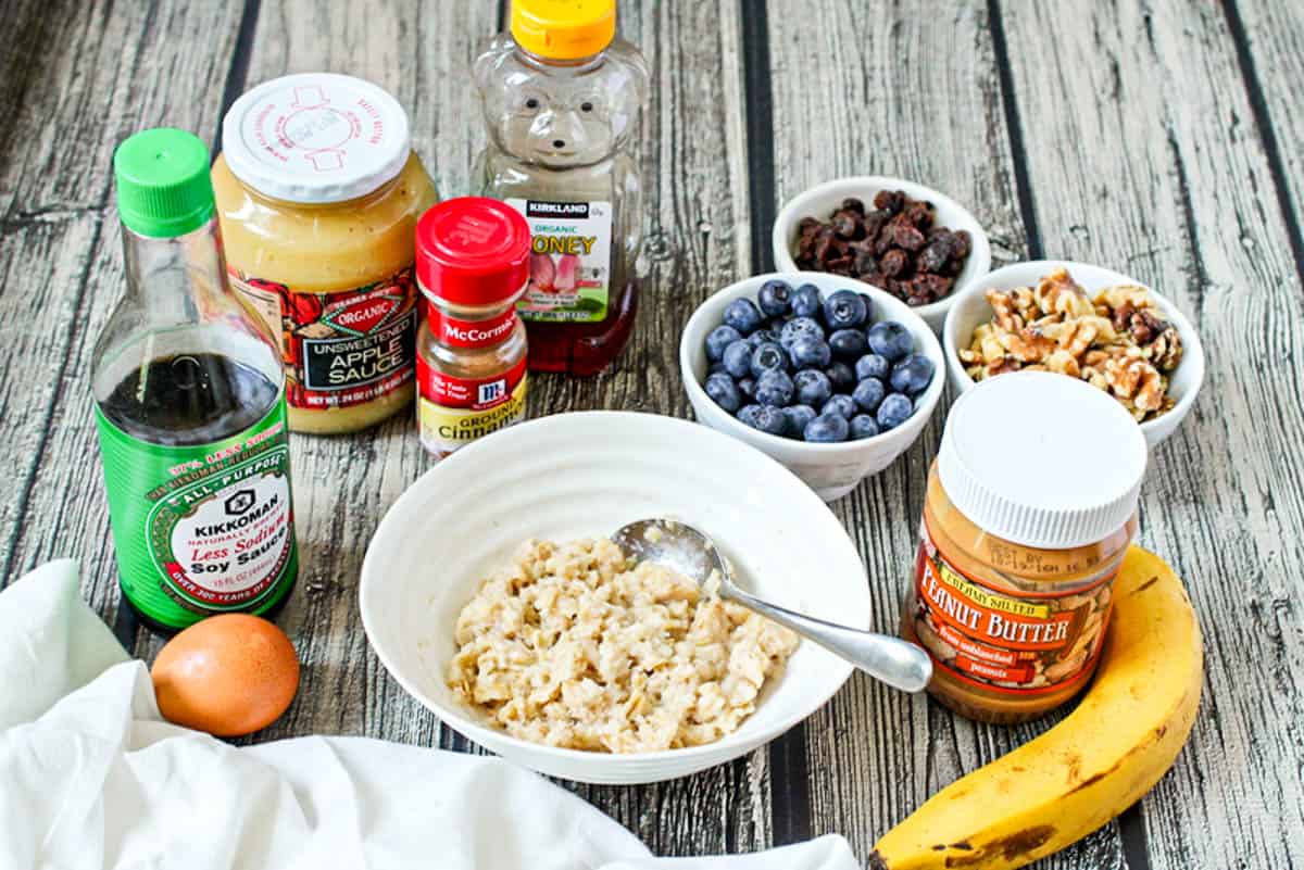 A bowl of oatmeal surrounded by toppings including blueberries, walnuts, applesauce, honey, soy sauce and an egg, peanut butter and banana.