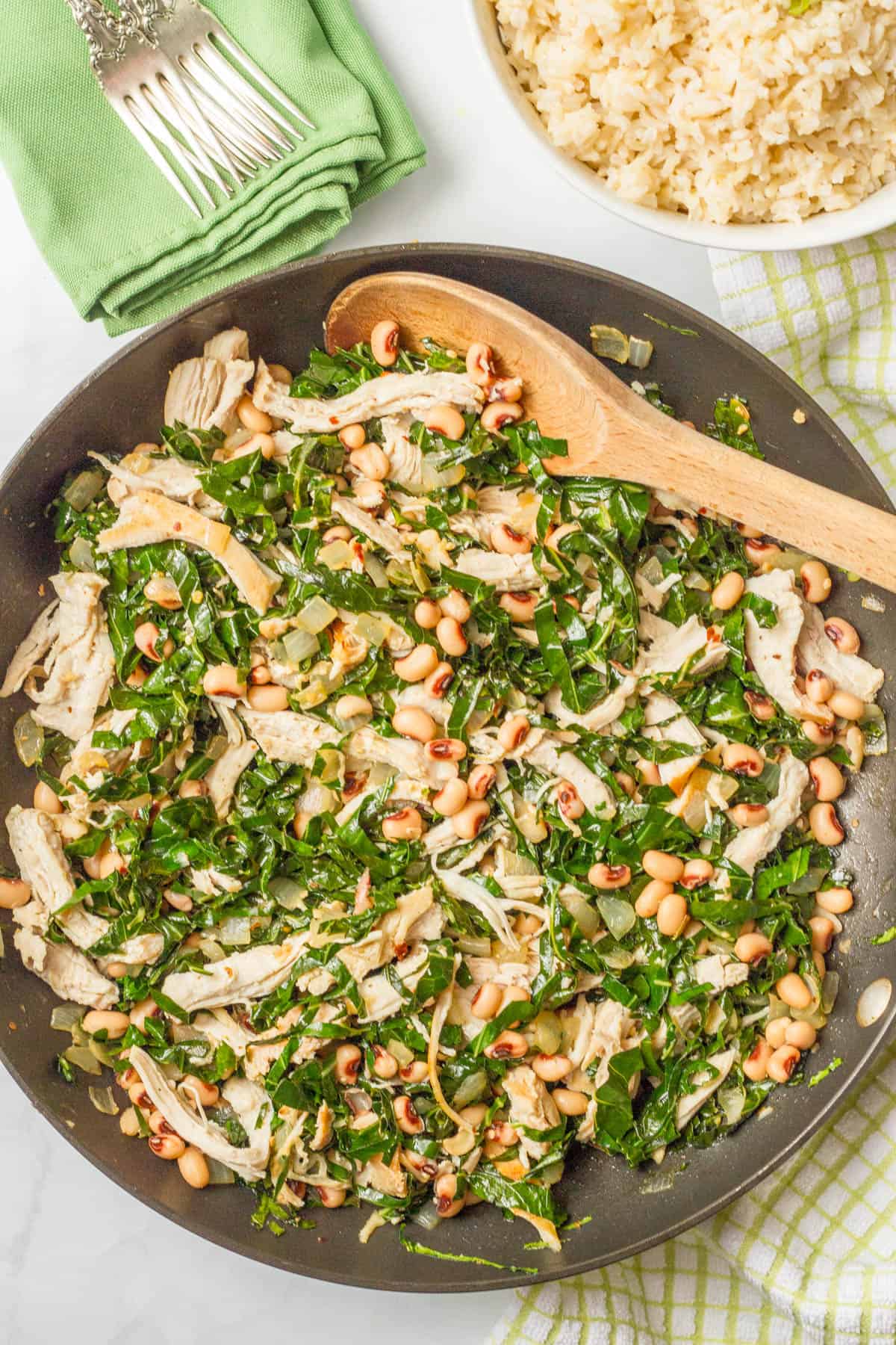 A dinner skillet with chicken, collards and black eyed peas with a wooden spoon resting in it and a bowl of rice to the side.