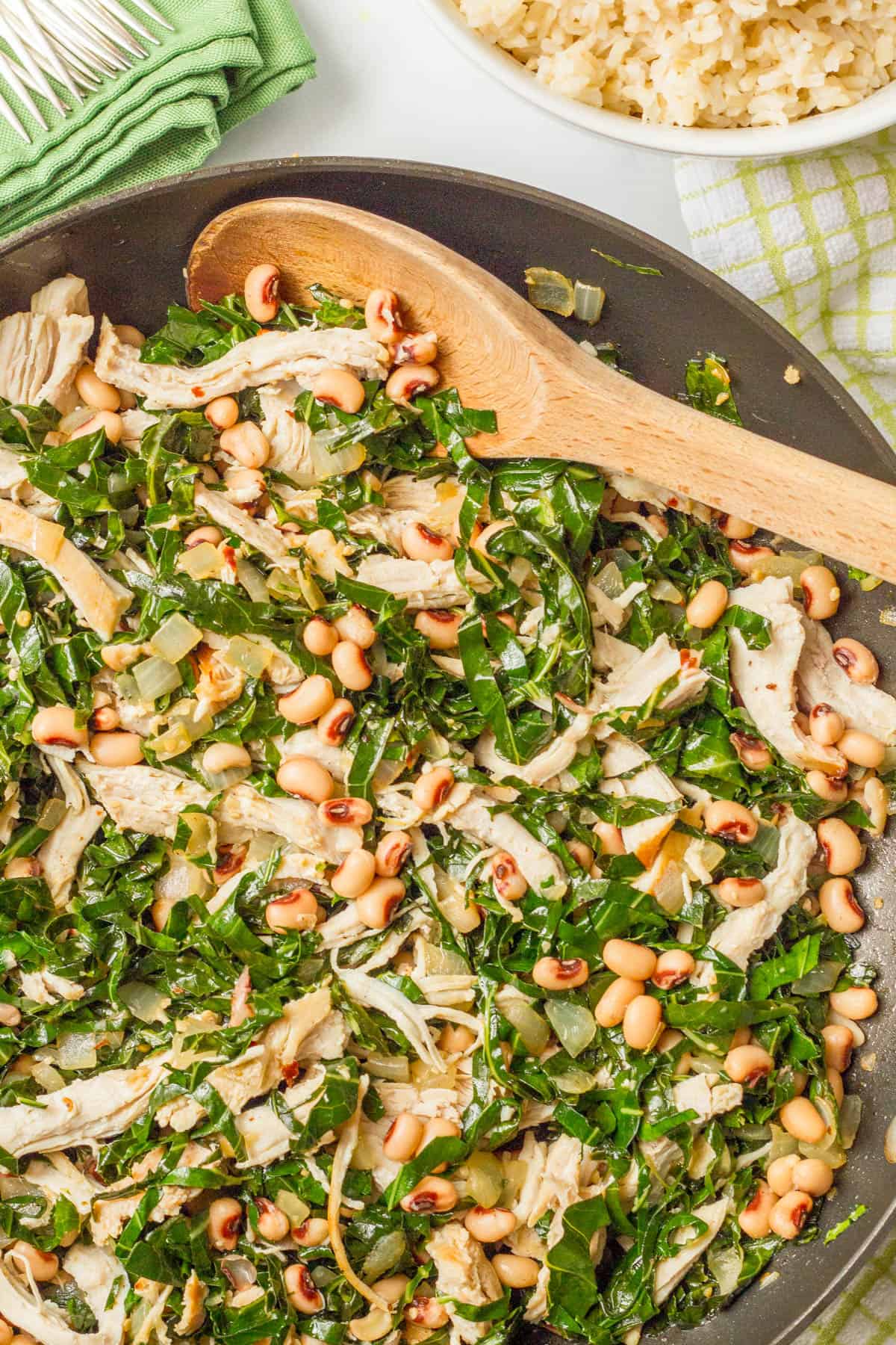 Close up of a dinner skillet with chicken, collards and black eyed peas with a wooden spoon resting in it and a bowl of rice to the side.