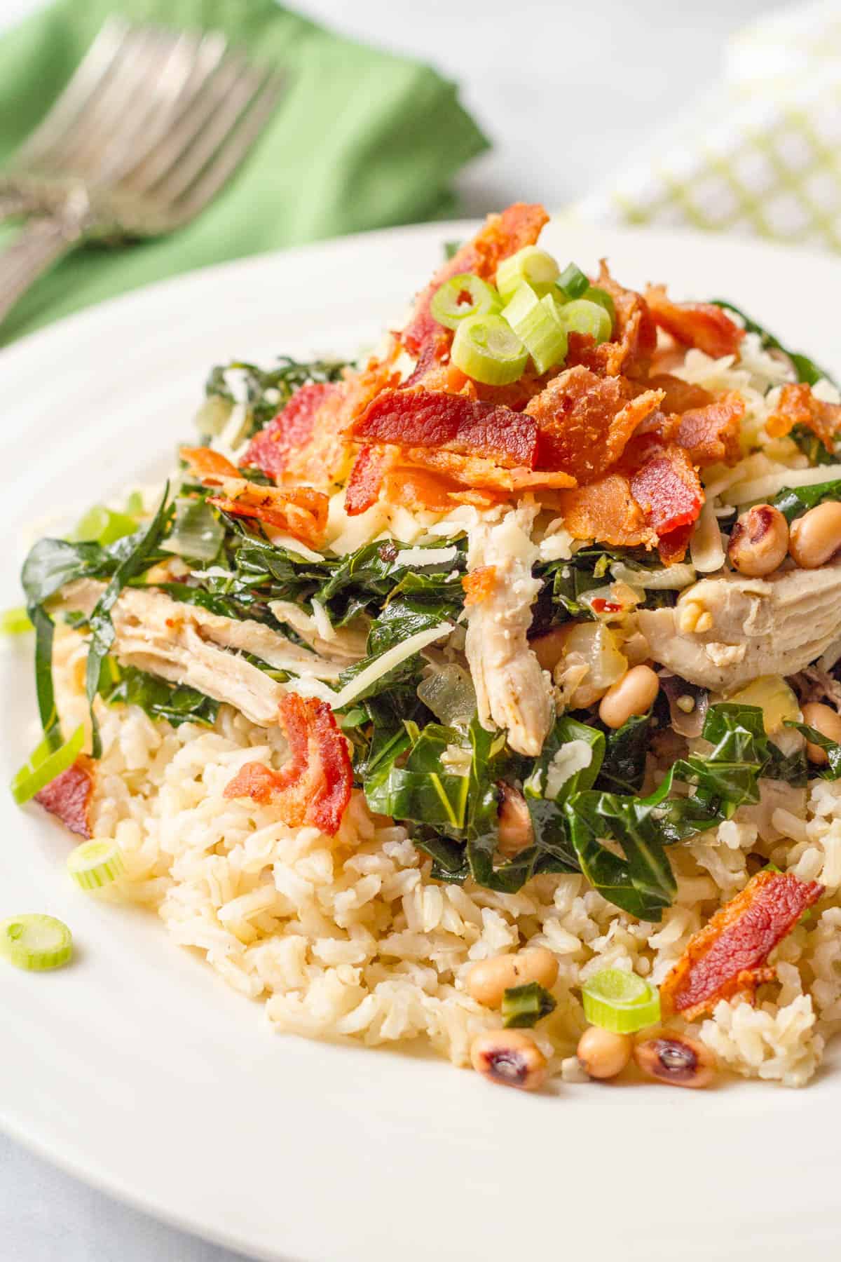 A plate with brown rice topped with a mixture of chicken, collards and black eyed peas with cooked, crumbled bacon and sliced green onions sprinkled on top.