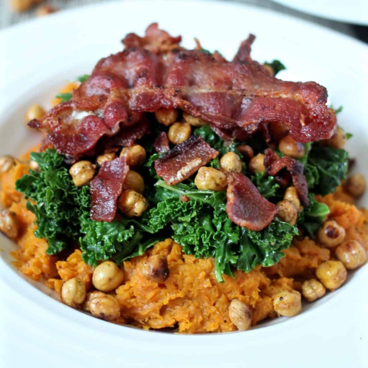 A low white bowl with mashed sweet potatoes topped with crispy roasted chickpeas, sautéed wilted kale and cooked, crumbled bacon.