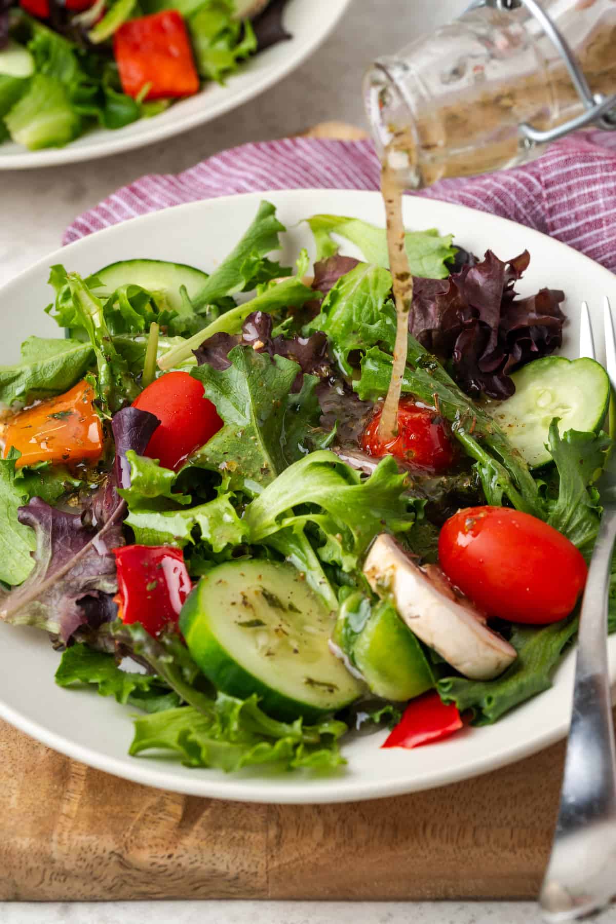 Italian dressing being poured from a jar over a salad of mixed greens with cucumber and tomato.