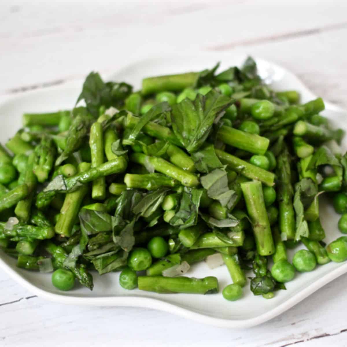 Close up of a fluted white plate with sautéed asparagus and green peas with strips of fresh basil.