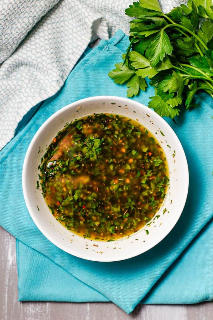 A white bowl with an herby chimichurri sauce set on teal napkins with fresh parsley to the side.