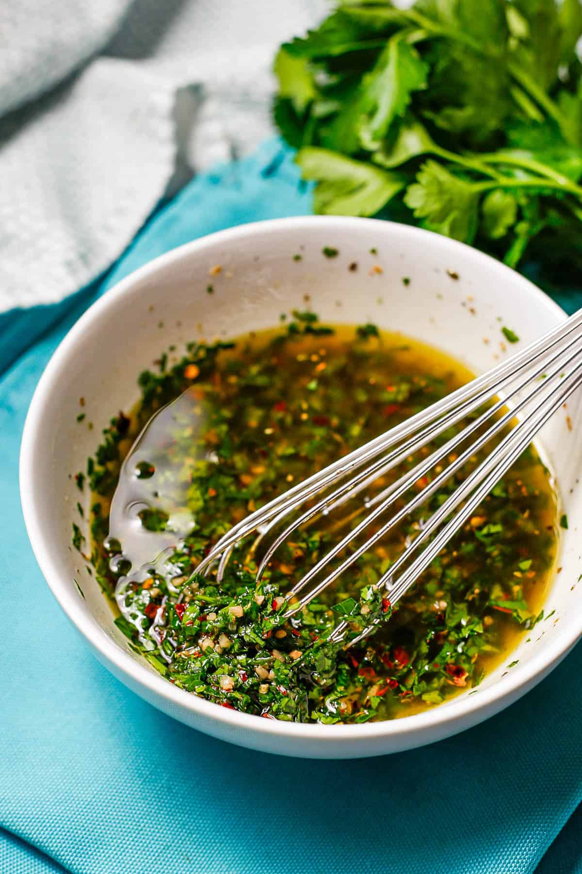 A whisk resting in a bowl of homemade chimichurri sauce.