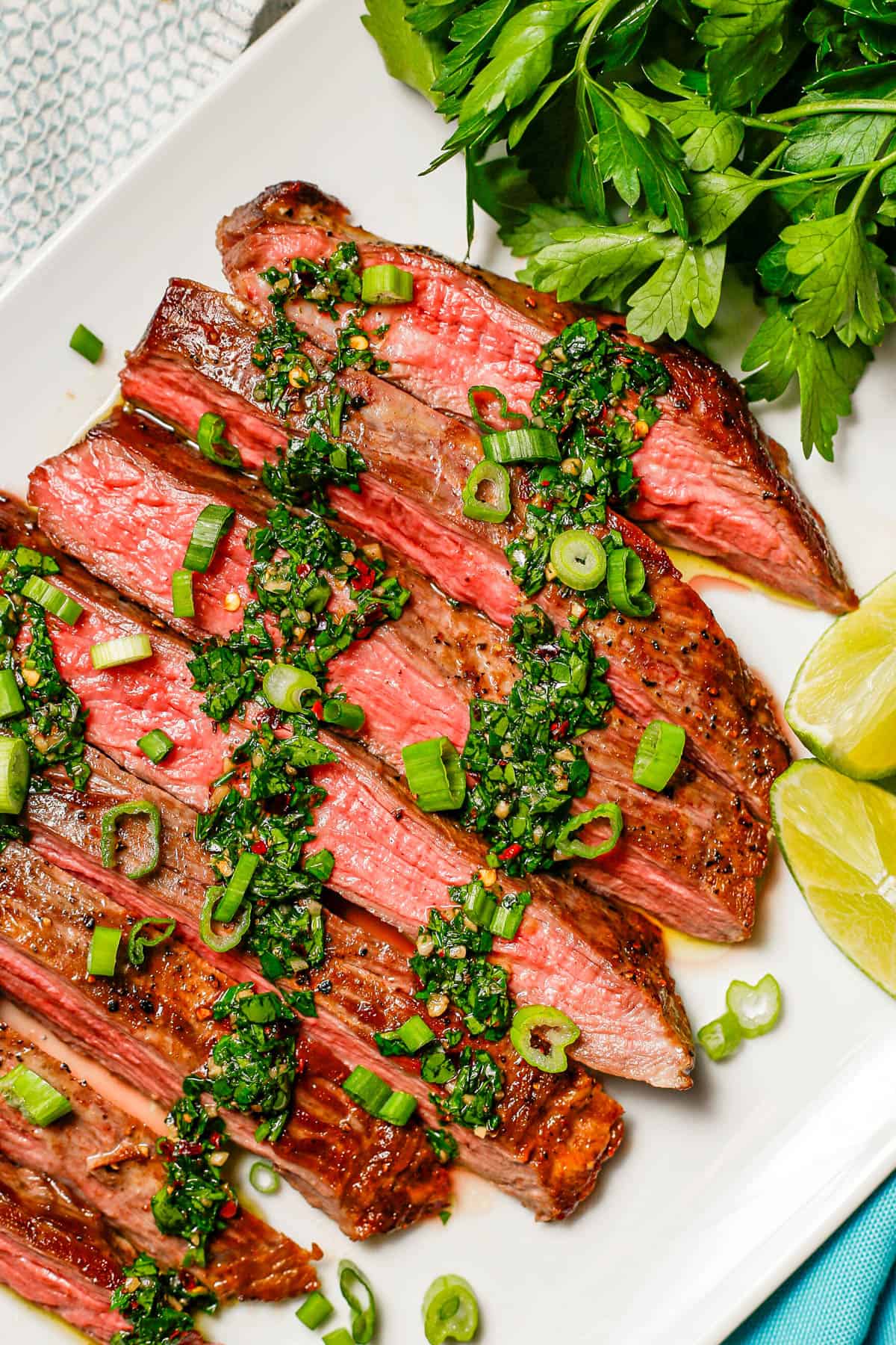 Close up of a white platter with sliced flank steak topped with chimichurri and sliced green onions with parsley and lime wedges to the side.