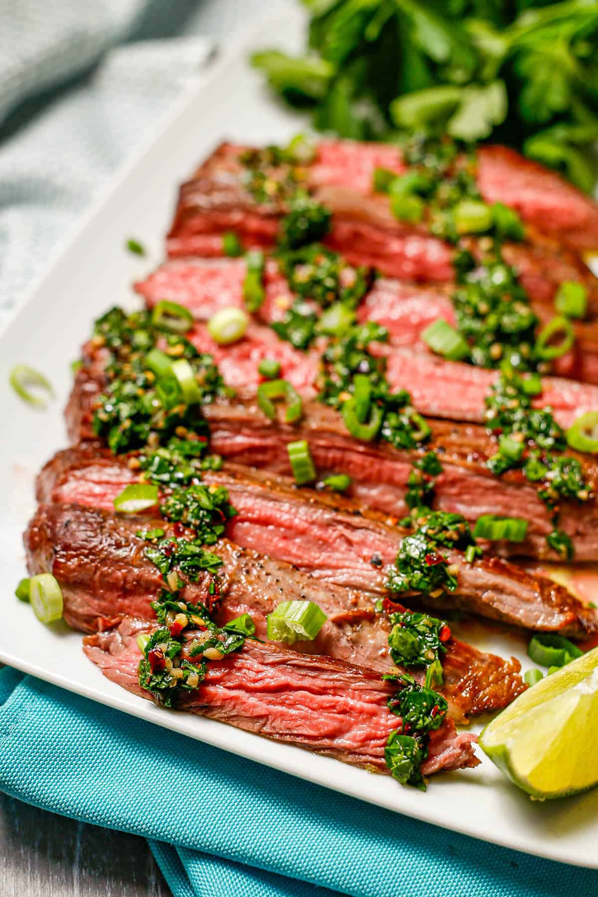 Sliced flank steak topped with homemade chimichurri sauce and sliced green onions on a white rectangular platter.